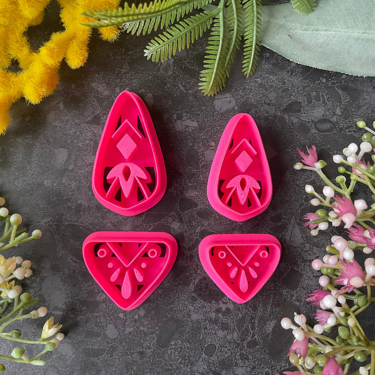 Bright pink 3d printed polymer clay cutters with floral and geometric designs are arranged on a dark textured surface with a black ruler at the bottom for scale, surrounded by decorative flowers and green leaves. Dune Drop Polymer Clay Cutter Set