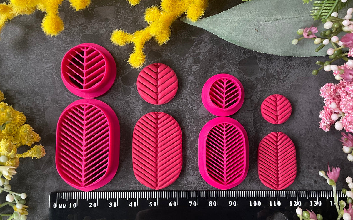 A set of bright pink 3d printed leaf-shaped polymer clay cutters arranged on a dark gray textured surface, surrounded by yellow and pink flowers. Etched Forms Polymer Clay Cutter Set
