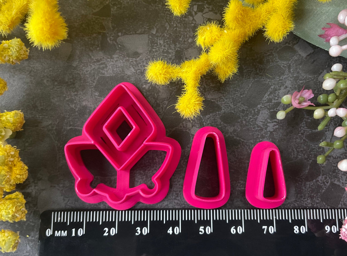 A set of bright pink and turquoise 3d printed polymer clay cutters arranged on a dark textured surface with decorative flowers around the edges. The set includes large fan-shaped stamping cutters, smaller matching cutters, and pointed cone-shaped cutters. First Light Polymer Clay Cutter Set