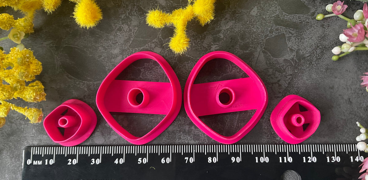 A flat-lay of polymer clay pieces and 3d printed polymer clay cutters on a dark gray textured surface, surrounded by decorative yellow, green, and pink flowers. The pieces include two bright magenta-pink geometric shaped polymer clay cutters and two light lavender-gray organic shapes, with smaller matching spacers. Organic Pebble Petal Polymer Clay Cutter Set