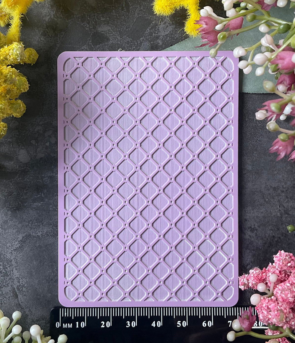 A rectangular lavender purple silicone mold with a raised geometric trellis pattern, displayed on a dark gray slate background and surrounded by yellow and pink flowers, with a ruler at the bottom showing its dimensions.