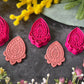 Three teardrop-shaped polymer clay cutters in hot pink and two shades of coral, arranged on a dark gray textured surface with decorative yellow and pink flowers. Afterglow Drop Polymer Clay Cutter