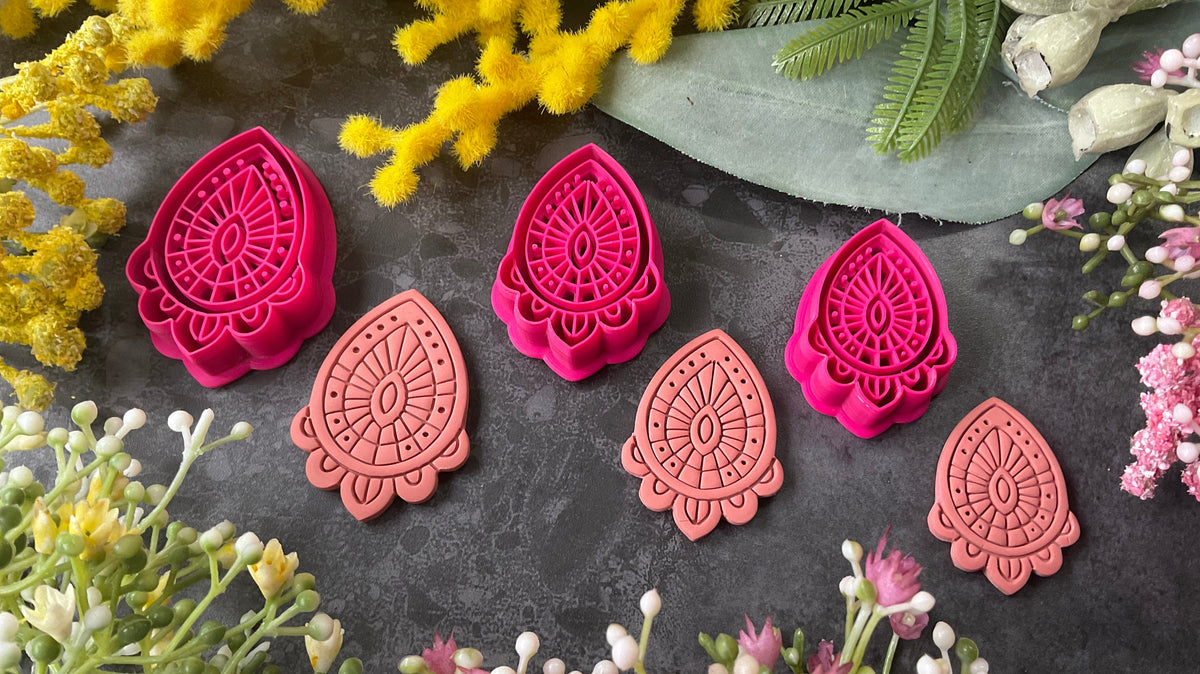 Three teardrop-shaped polymer clay cutters in hot pink and two shades of coral, arranged on a dark gray textured surface with decorative yellow and pink flowers. Afterglow Drop Polymer Clay Cutter