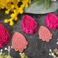Three teardrop-shaped polymer clay cutters in hot pink and two shades of coral, arranged on a dark gray textured surface with decorative yellow and pink flowers. Afterglow Drop Polymer Clay Cutter