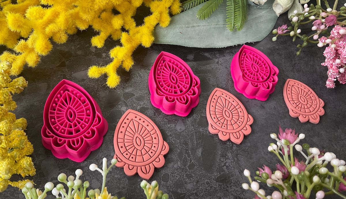 Three teardrop-shaped polymer clay cutters in hot pink and two shades of coral, arranged on a dark gray textured surface with decorative yellow and pink flowers. Afterglow Drop Polymer Clay Cutter