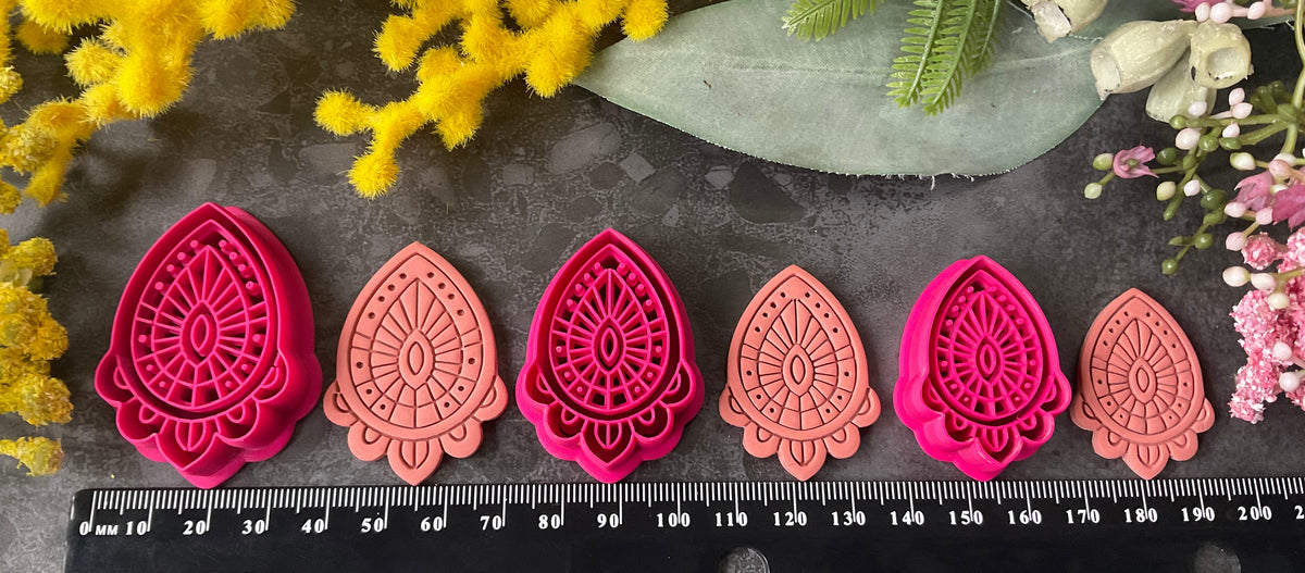 Three teardrop-shaped polymer clay cutters in hot pink and two shades of coral, arranged on a dark gray textured surface with decorative yellow and pink flowers. Afterglow Drop Polymer Clay Cutter