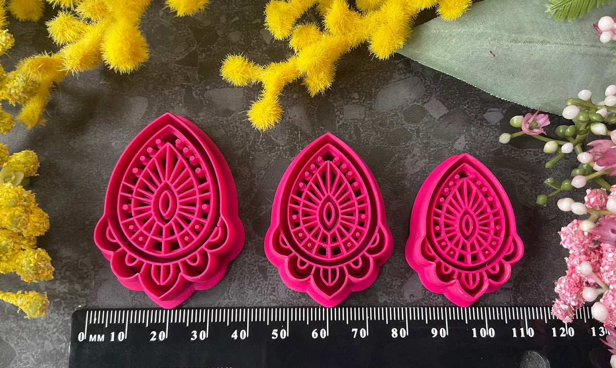 Three teardrop-shaped polymer clay cutters in hot pink and two shades of coral, arranged on a dark gray textured surface with decorative yellow and pink flowers. Afterglow Drop Polymer Clay Cutter
