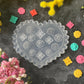 A heart-shaped clear plastic mold with embossed floral patterns, surrounded by colorful small flower-shaped pieces and yellow and pink flowers on a dark textured surface.