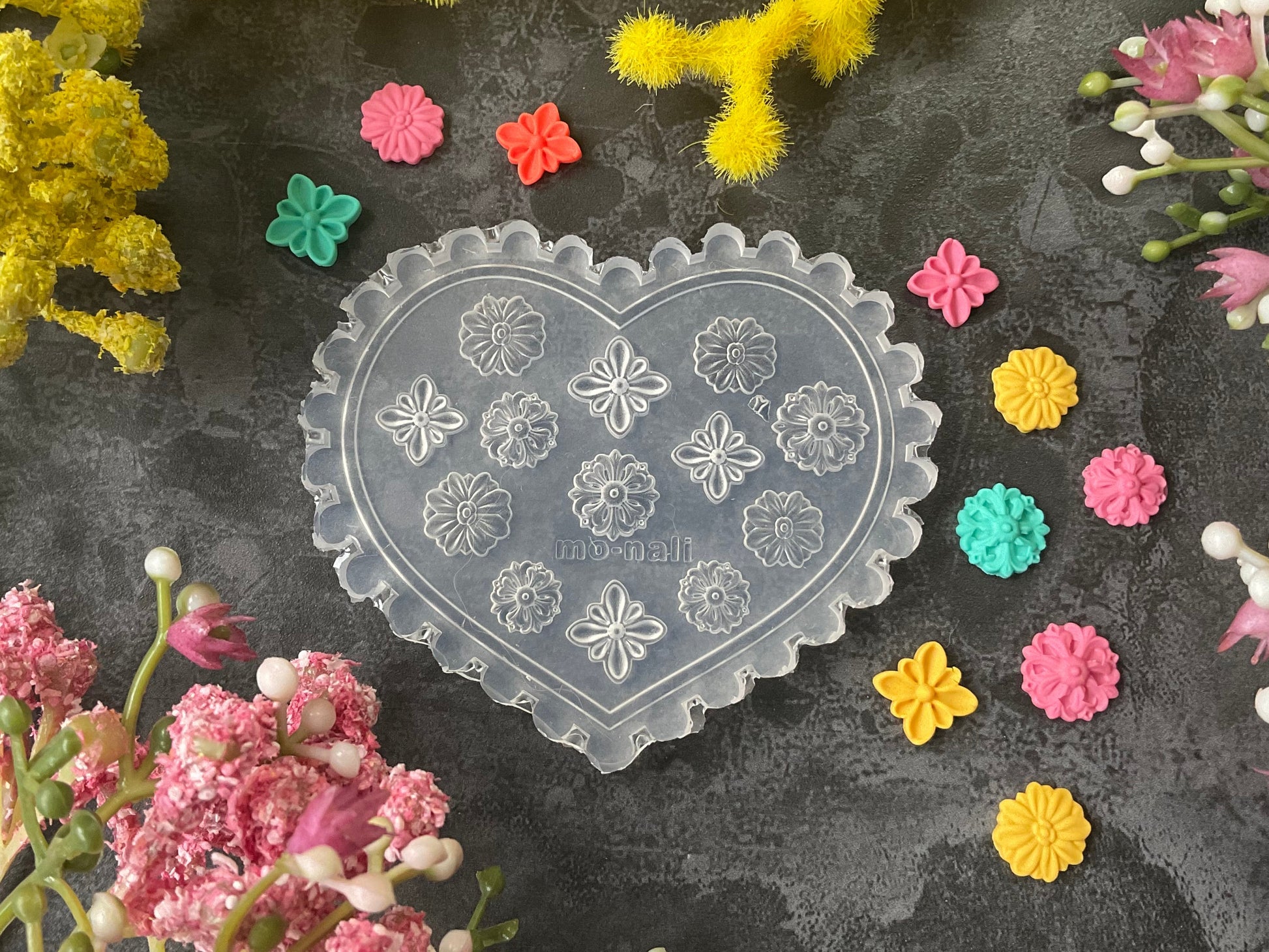 A heart-shaped clear plastic mold with embossed floral patterns, surrounded by colorful small flower-shaped pieces and yellow and pink flowers on a dark textured surface.