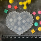 A heart-shaped clear plastic stamp or mold with embossed snowflake and floral patterns, displayed on a dark textured surface with a black ruler at the bottom for scale. The scene is styled with small colorful flower stamps in pink, yellow, green, and orange, surrounded by yellow and pink flowers.