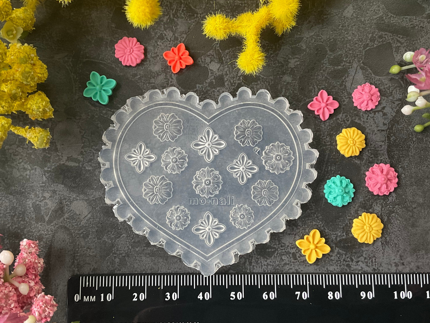 A heart-shaped clear plastic stamp or mold with embossed snowflake and floral patterns, displayed on a dark textured surface with a black ruler at the bottom for scale. The scene is styled with small colorful flower stamps in pink, yellow, green, and orange, surrounded by yellow and pink flowers.