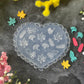 A heart-shaped clear plastic mold with embossed autumn leaf designs, displayed on a dark textured surface surrounded by colorful artificial flowers.