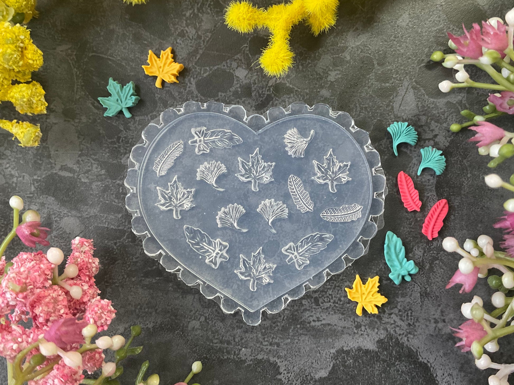 A heart-shaped clear plastic mold with embossed autumn leaf designs, displayed on a dark textured surface surrounded by colorful artificial flowers.