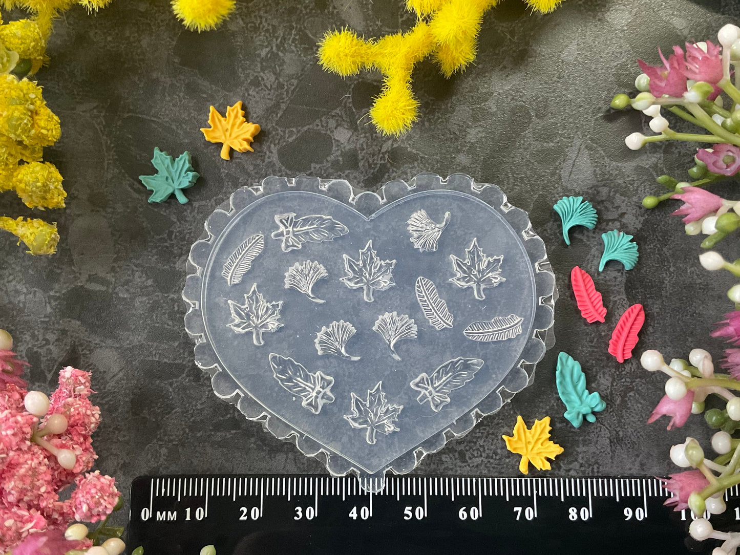 A heart-shaped clear plastic mold with embossed leaf patterns, displayed on a dark textured surface with a black ruler for scale and surrounded by colorful faux flowers.