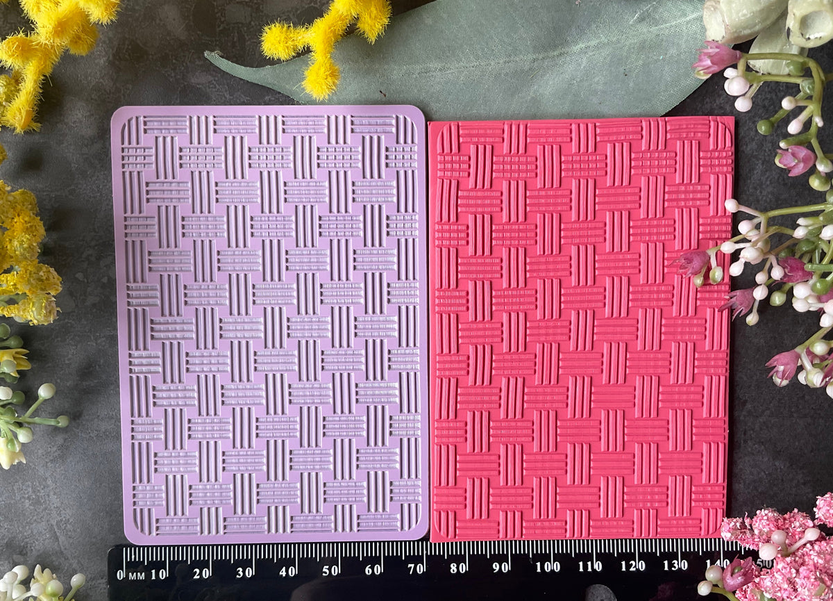 Two textured rectangular cards or texture mats in a bamboo weave pattern, one light lavender and one coral pink, displayed side-by-side on a dark surface with a black measuring ruler at the bottom and yellow and pink flowers framing the scene.