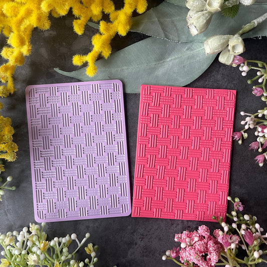 Two rectangular textured cards or stamps in lavender purple and bright pink in a bamboo weave pattern, arranged on a dark surface with yellow and pink flowers around them