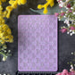 A lavender purple rectangular silicone mold with a bamboo basketweave pattern, displayed on a dark gray surface with a black ruler underneath for scale, surrounded by yellow and pink flowers.
