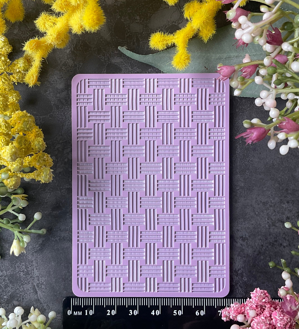A lavender purple rectangular silicone mold with a bamboo basketweave pattern, displayed on a dark gray surface with a black ruler underneath for scale, surrounded by yellow and pink flowers.