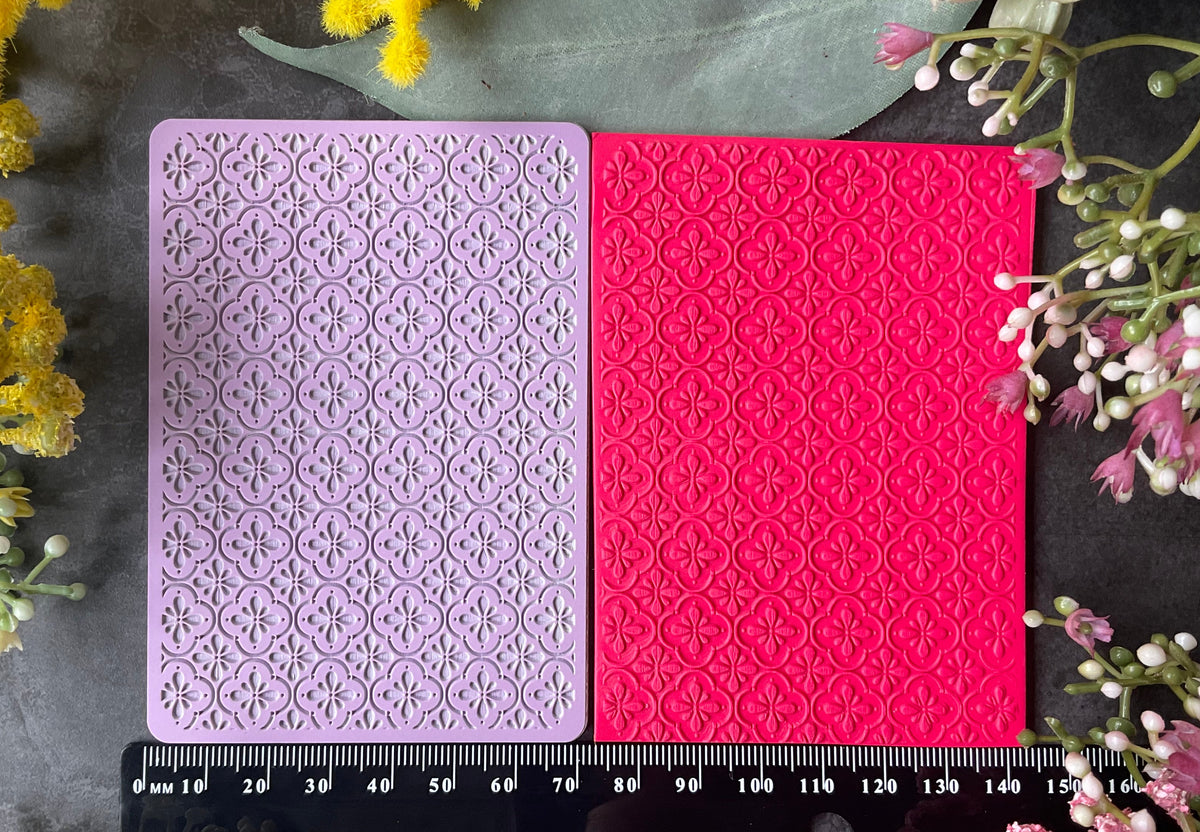 Two rectangular craft sheets with embossed Mediteranean floral tile patterns, one lavender and one bright pink, displayed on a dark surface with a black ruler at the bottom and decorative flowers around the edges.