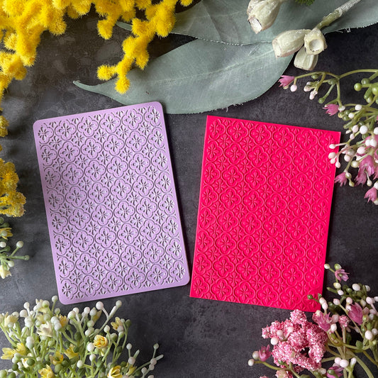 Two rectangular embossed cardstock sheets, one lavender and one bright pink with a Mediterranean floral tile pattern, arranged on a dark textured surface with yellow and pink flowers around them.