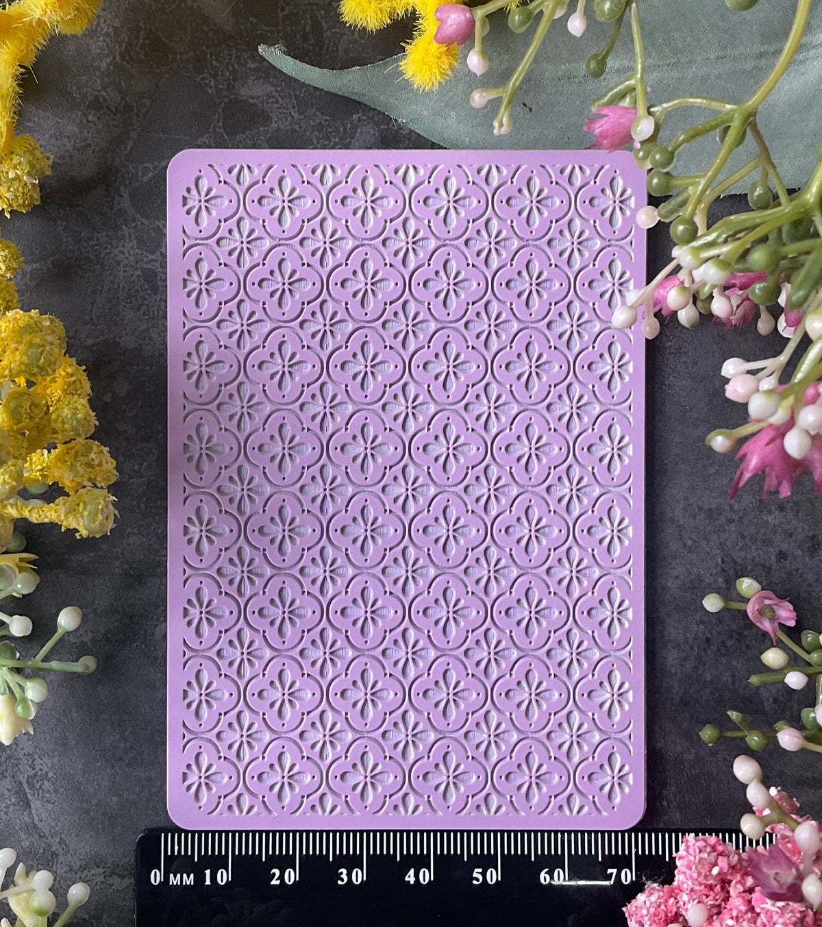 A lavender purple rectangular embossed stamp or plaque with a repeating Mediteranean floral trellis tile pattern, displayed on a dark textured surface and surrounded by yellow and pink flowers, with a black ruler at the bottom showing measurements.