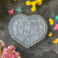 A heart-shaped clear plastic mold with butterfly and floral embossed designs, styled on a dark gray textured surface surrounded by yellow and pink flowers.