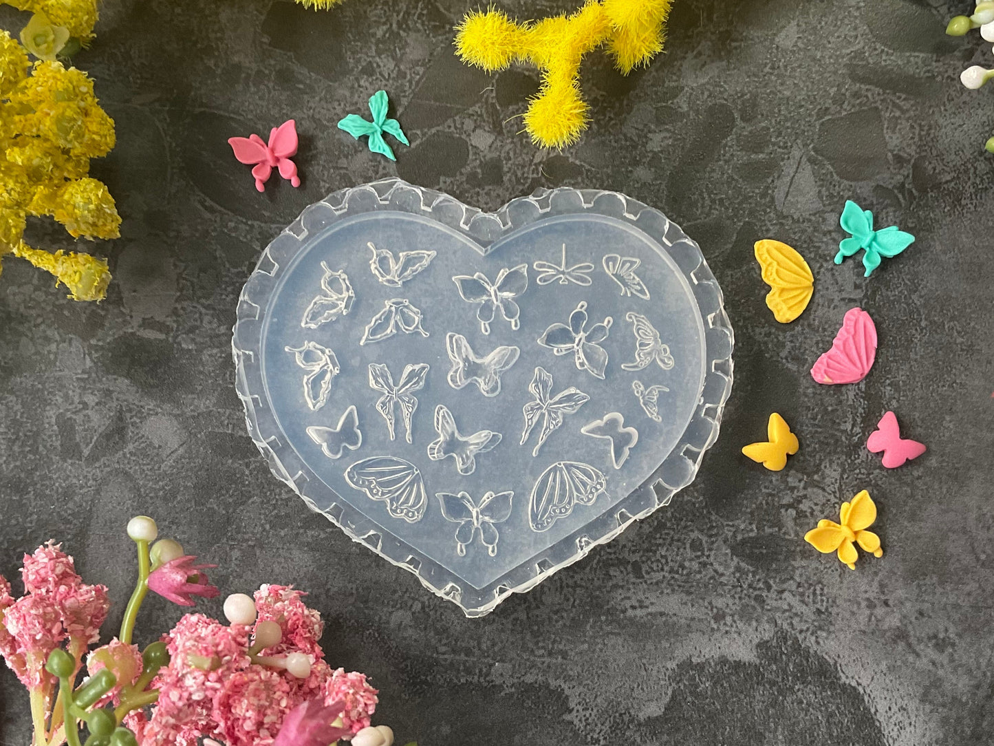 A heart-shaped clear plastic mold with butterfly and floral embossed designs, styled on a dark gray textured surface surrounded by yellow and pink flowers.