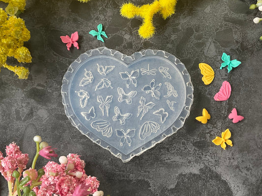 A heart-shaped clear plastic mold with butterfly and floral embossed designs, styled on a dark gray textured surface surrounded by yellow and pink flowers.