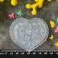 A heart-shaped translucent plastic mold with butterfly and floral engravings, displayed on a dark textured surface with a black ruler at the bottom for scale, surrounded by yellow and pink flowers and small colorful butterfly decorations.