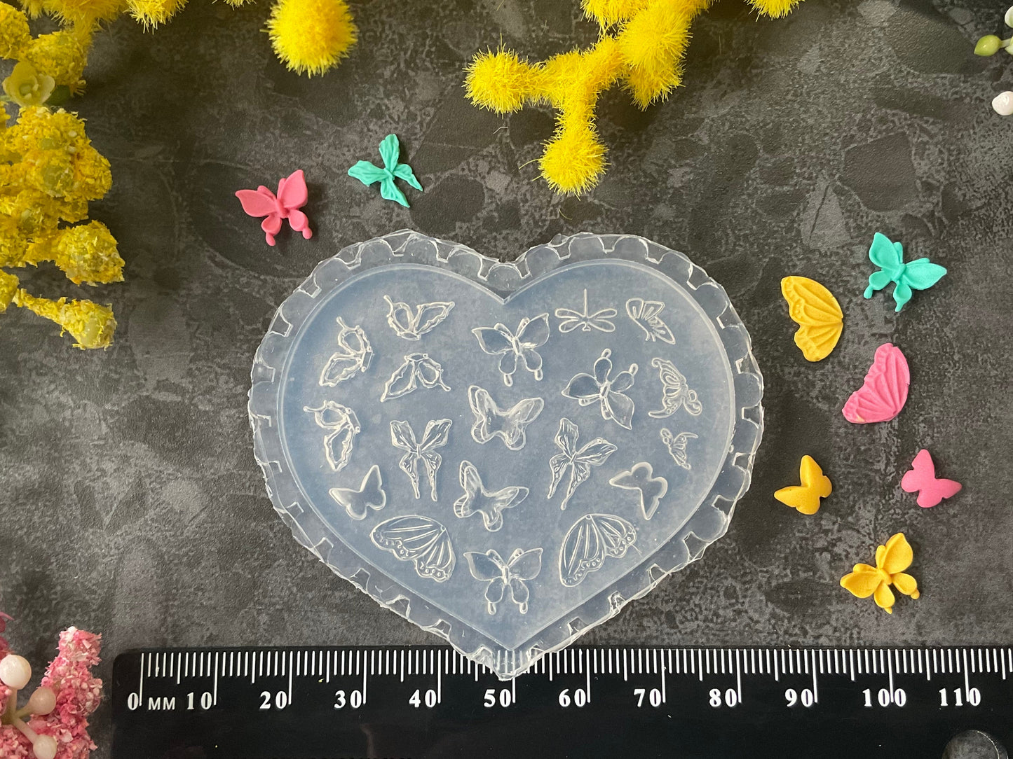 A heart-shaped translucent plastic mold with butterfly and floral engravings, displayed on a dark textured surface with a black ruler at the bottom for scale, surrounded by yellow and pink flowers and small colorful butterfly decorations.