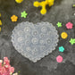 A heart-shaped clear plastic craft mold with floral engravings, displayed on a dark textured surface surrounded by colorful artificial flowers.