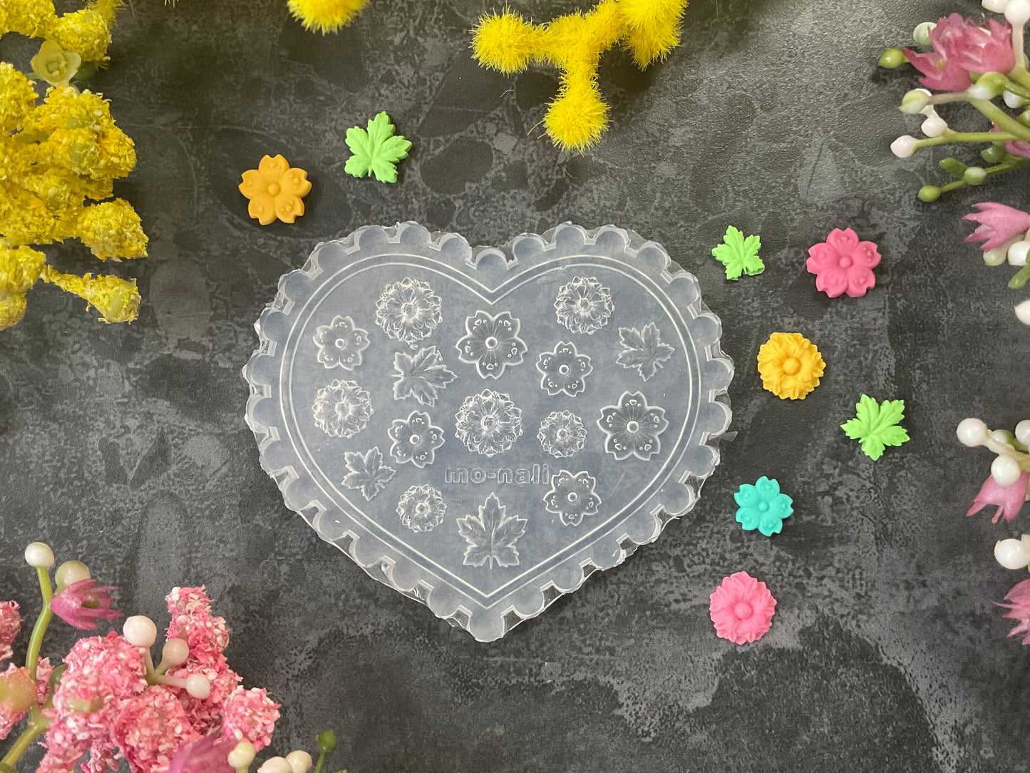 A heart-shaped clear plastic craft mold with floral engravings, displayed on a dark textured surface surrounded by colorful artificial flowers.