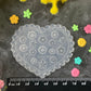 A heart-shaped clear plastic stamp or mold with floral engravings, displayed on a dark textured surface with a black ruler at the bottom for scale, surrounded by small colorful flower petals and faux flowers.