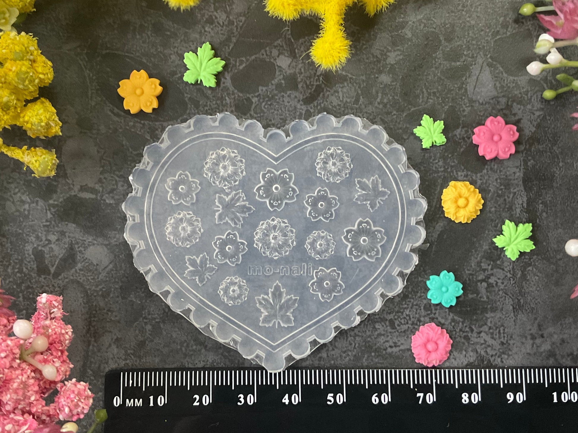 A heart-shaped clear plastic stamp or mold with floral engravings, displayed on a dark textured surface with a black ruler at the bottom for scale, surrounded by small colorful flower petals and faux flowers.