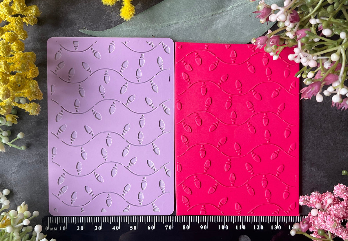 Two rectangular embossed stamping plates, one lavender and one bright pink with a Christmas fairy lights pattern, displayed on a dark textured surface with a black ruler at the bottom and yellow and pink flowers framing the scene.