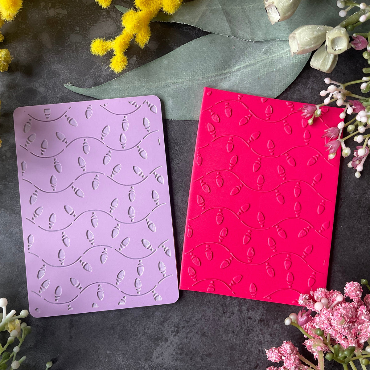 Two rectangular craft cards, one lavender purple and one bright pink, both embossed with a wavy Christmas fairy lights pattern, displayed on a dark gray textured surface surrounded by yellow and pink flowers.