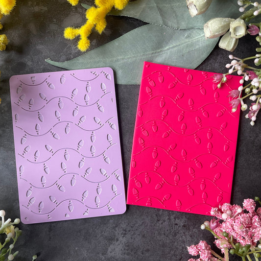 Two rectangular craft cards, one lavender purple and one bright pink, both embossed with a wavy Christmas fairy lights pattern, displayed on a dark gray textured surface surrounded by yellow and pink flowers.