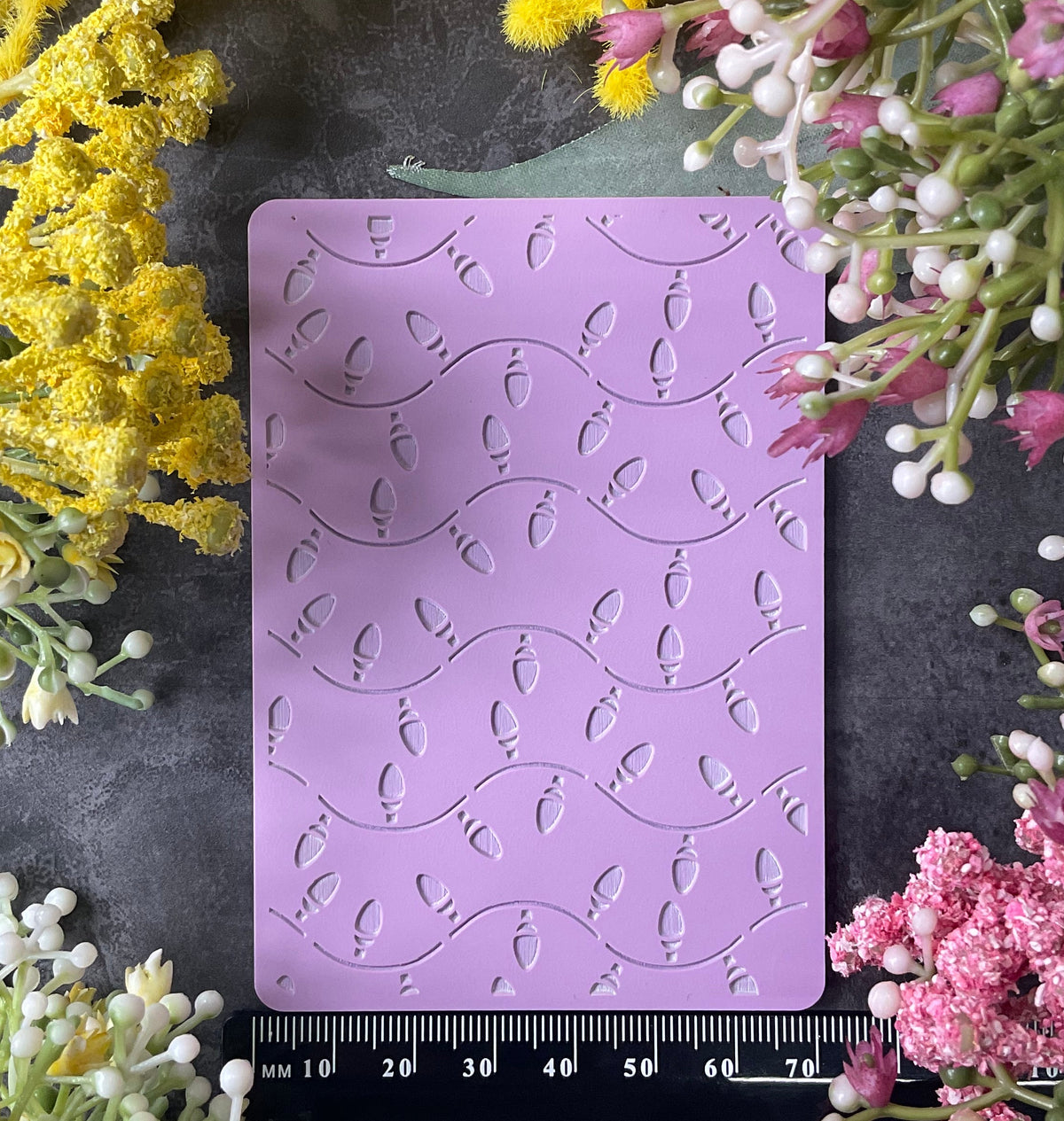 A lavender rectangular silicone stamp or mold with embossed Christmas fairy lights pattern, displayed on a dark textured surface and surrounded by yellow and pink flowers. A black ruler is visible along the bottom edge.