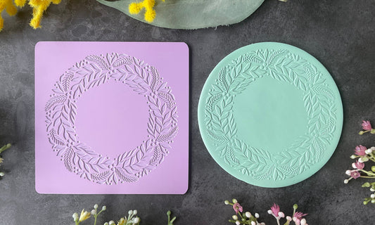 A lavender rectangular silicone mold with a Christmas Wreath Trinket Sized Texture Mat leafy wreath design and a mint green circular clay sample showing the pressed impression, styled with flowers on a dark gray textured background.