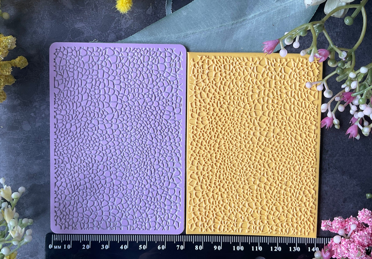 Two rectangular textured craft mats, one lavender purple and one mustard yellow Debossed Leather Texture Mat pattern, displayed side-by-side on a dark slate background with a black measuring ruler at the bottom and decorative flowers around the edges.