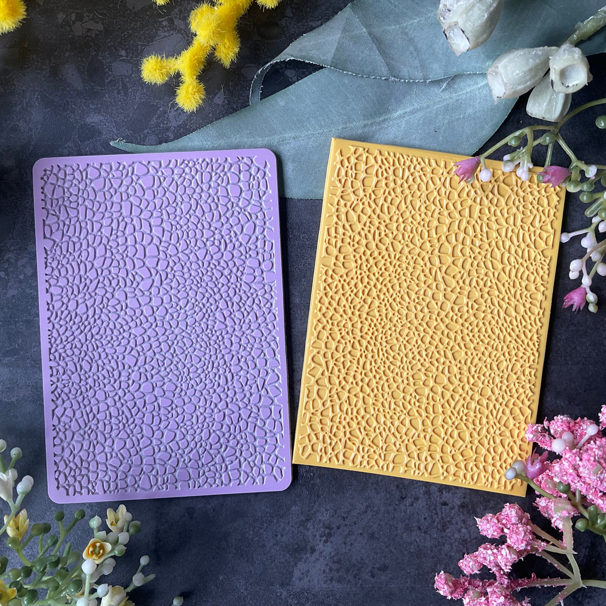 Two textured rectangular cards or stamps, one lavender purple and one mustard yellow Debossed Leather pattern Texture Mat displayed on a dark gray surface surrounded by decorative flowers and a blue ribbon.