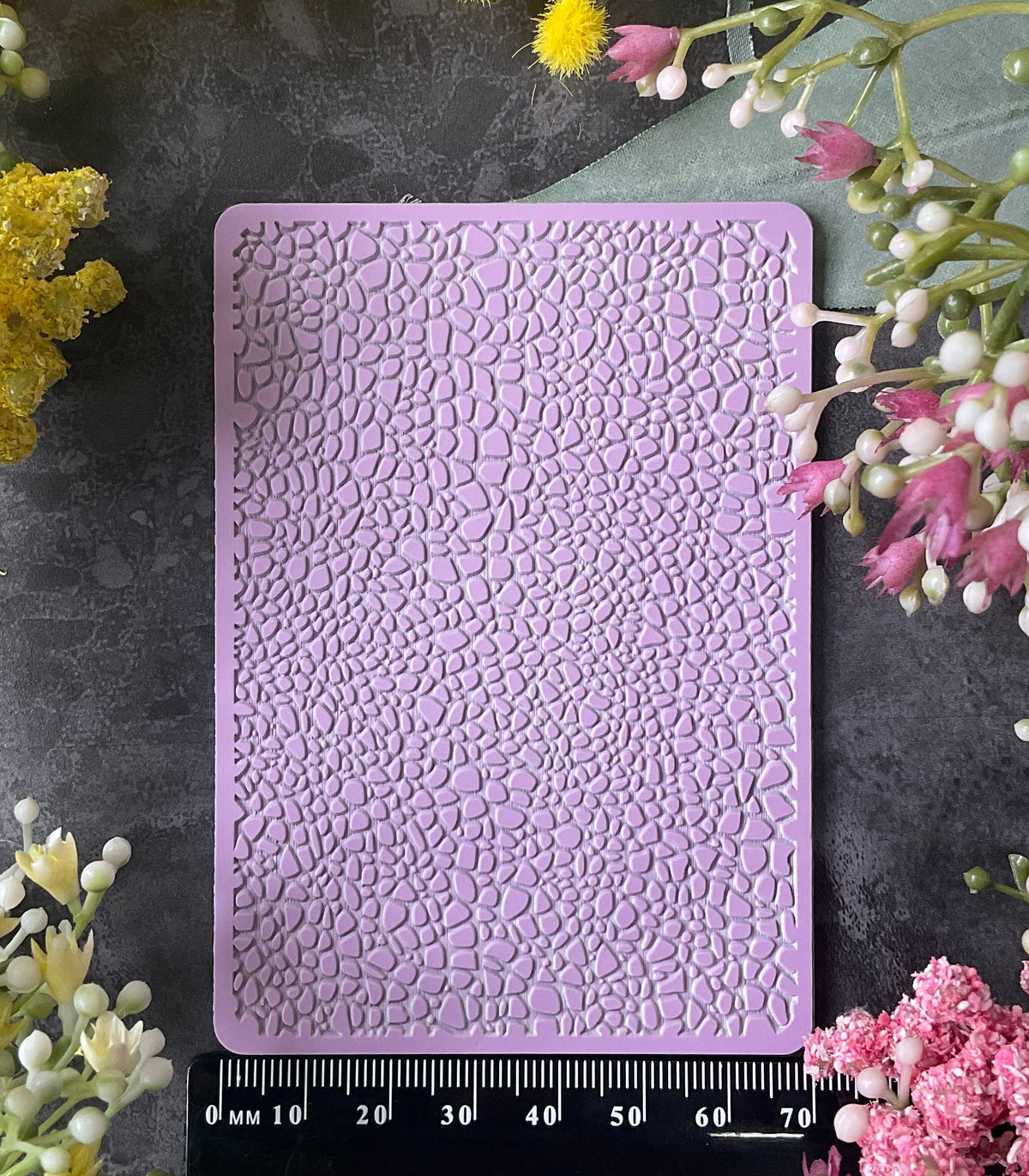 A lavender purple silicone mold with a Debossed Leather Texture Mat textured, cracked surface pattern, shown flat-lay on a dark gray background surrounded by yellow and pink flowers, with a black ruler at the bottom indicating its 70mm size.