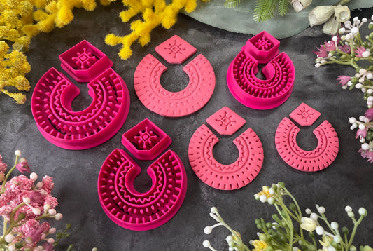 Three sets of 3d printed bright pink polymer clay cutters in different sizes arranged on a dark textured surface, surrounded by decorative yellow and white flowers. Desert Arch Polymer Clay Cutter Set