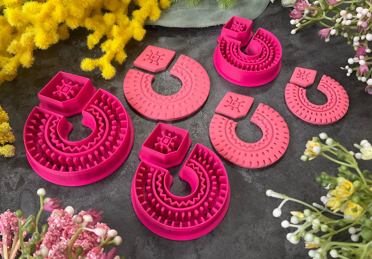 Three sets of 3d printed bright pink polymer clay cutters in different sizes arranged on a dark textured surface, surrounded by decorative yellow and white flowers. Desert Arch Polymer Clay Cutter Set