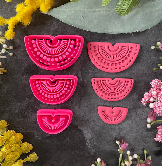 Six bright pink 3d printed half-moon polymer clay cutters in various sizes, arranged on a dark textured surface surrounded by yellow, green, and pink flowers. Desert Rainbow Halo Polymer Clay Cutter Set