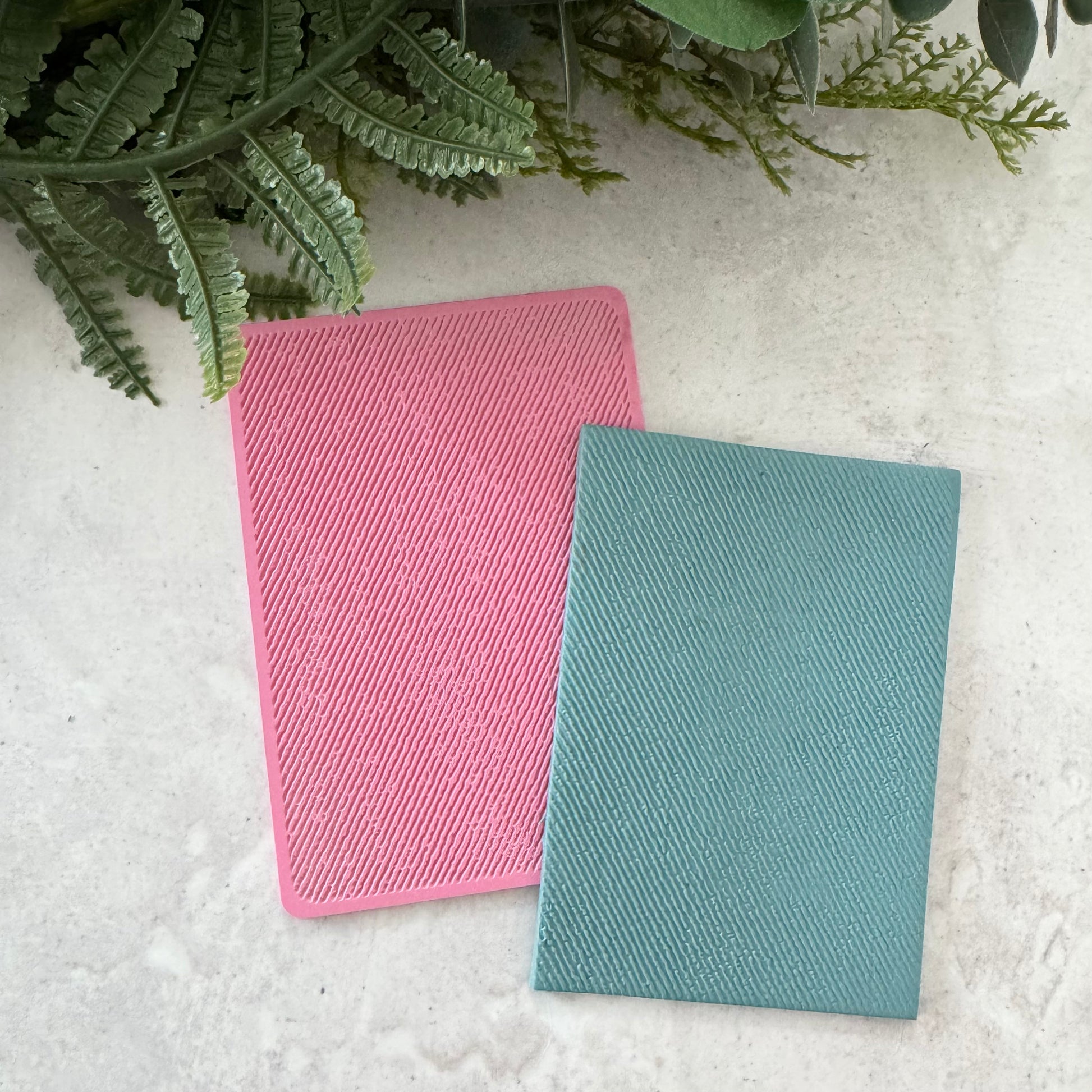 Two textured rectangular cards or sleeves, one pink and one teal, arranged on a light stone surface with green fern leaves at the top.