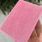 Pink textured square object held by a hand with a natural background