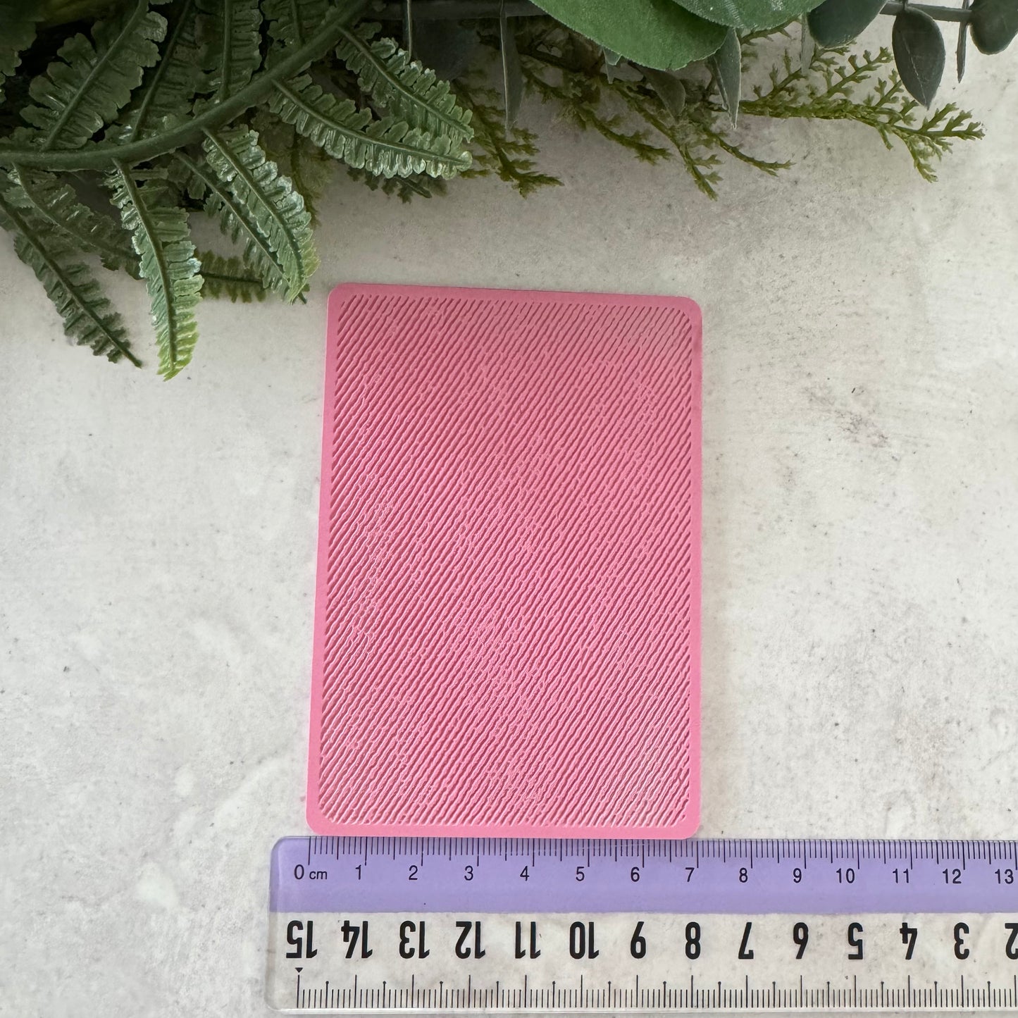A pink rectangular silicone mold with a textured, ribbed surface, shown on a light gray stone background with a purple ruler at the bottom for scale and green foliage at the top.