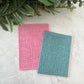 Two textured rectangular cards or mats, one pink and one teal, arranged on a light stone surface with green foliage at the top.