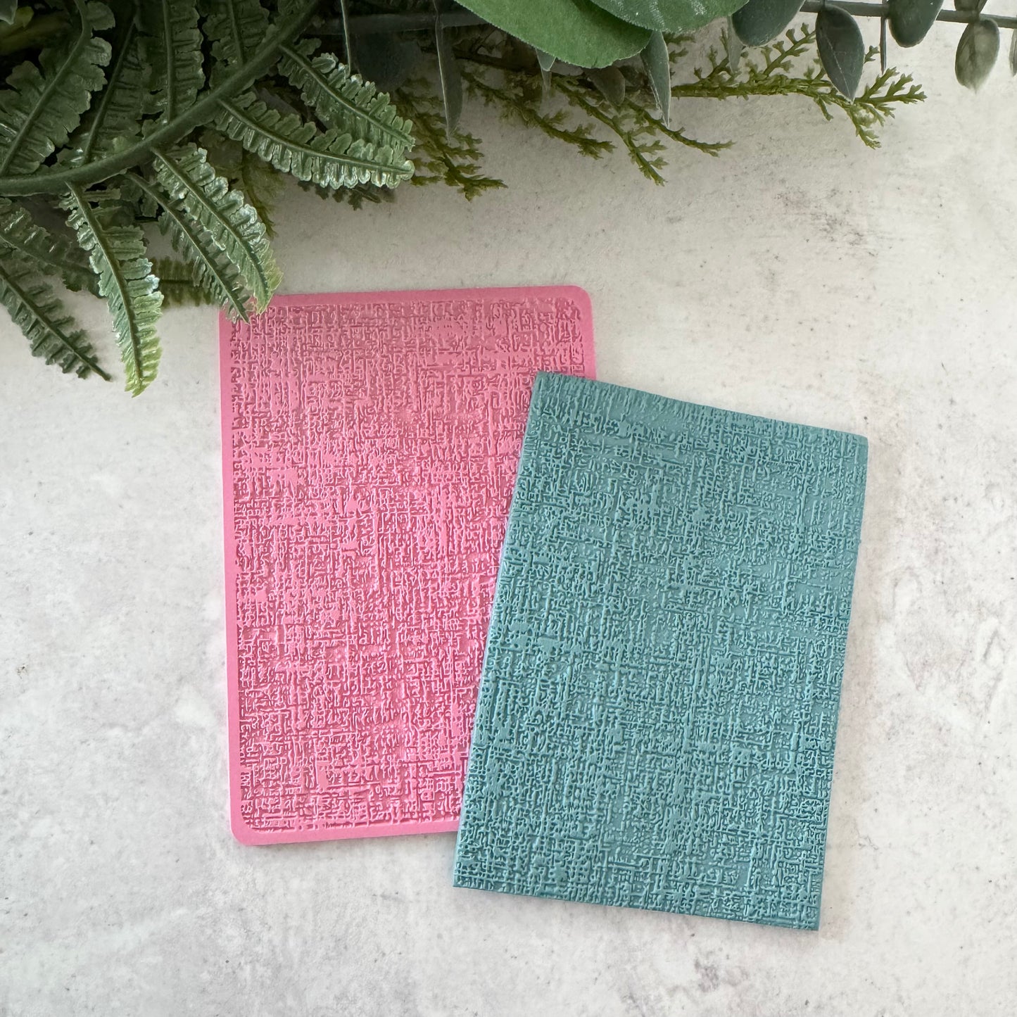 Two textured rectangular cards or mats, one pink and one teal, arranged on a light stone surface with green foliage at the top.
